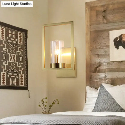 Modern brass-framed wall light with clear glass cylinder housing an exposed bulb for contemporary bedroom use
