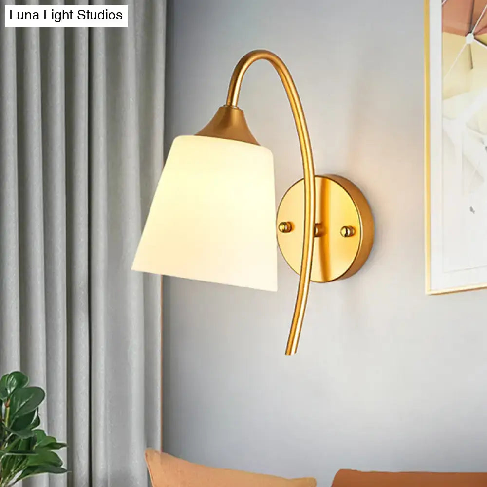 Contemporary gold milk glass wall lamp with a gold-toned curved arm and white tapered glass shade for bedroom use