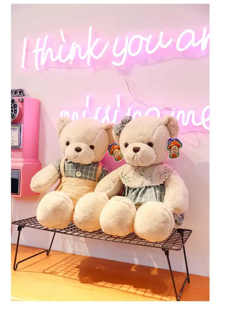 Two plush toys couple bear plush dressed in overalls and lace-trimmed dress sitting on a wire rack by decorbites