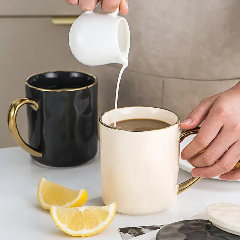Cream-colored ceramic gold rim mug being filled with milk from white pitcher part of nordic gold rim rim mug set