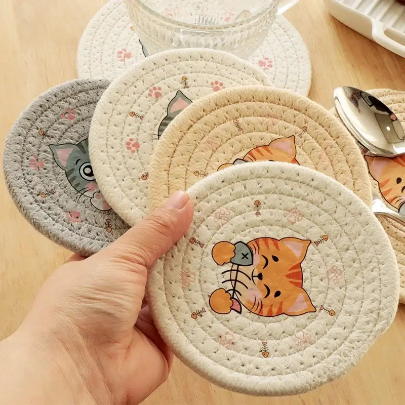 A hand holds a cream braided rope cat print round coaster with an orange tabby design and pink paw prints