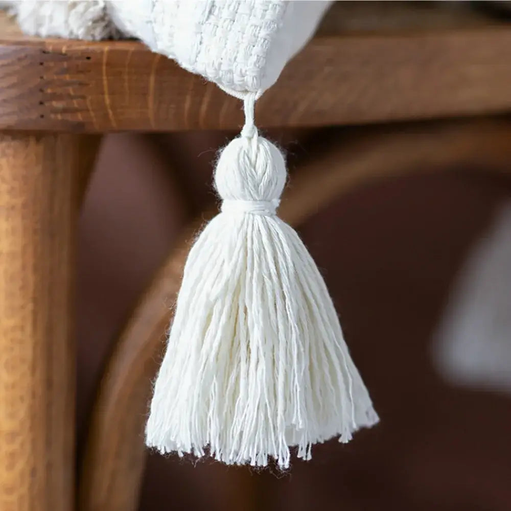 A cream geometric embroidery cushion cover with textured loosely woven cotton tassels hanging from a wooden surface
