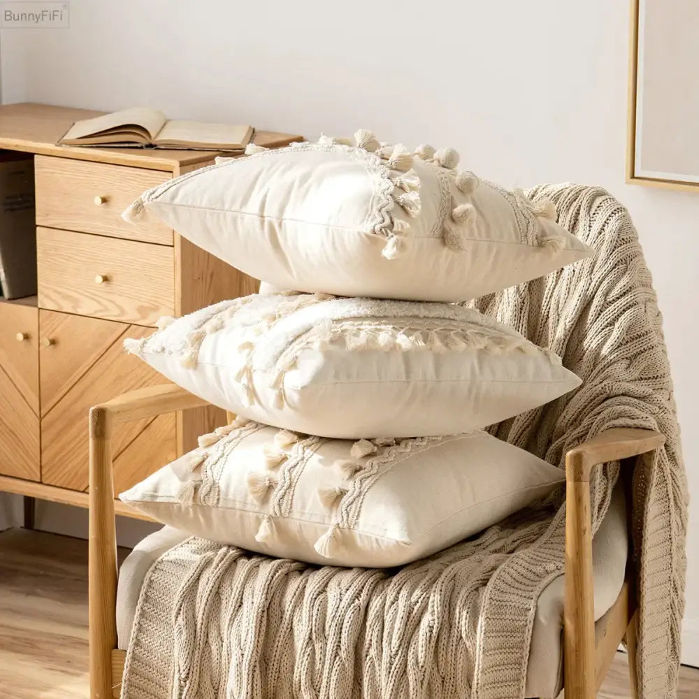 Stack of three cream ivory pompom tassel pillow covers with textured embroidery and tassel trim on wooden chair decorbites ivory pompom