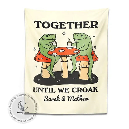 Cream-colored wall tapestry featuring a cute cartoon frog moon phase design with two frogs on red mushrooms sipping coffee