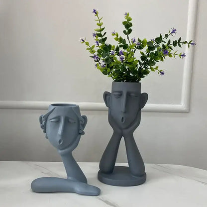 Gray ceramic face-shaped flowerpot ornament with matte finish holding greenery and purple flowers by decorbites abstract family
