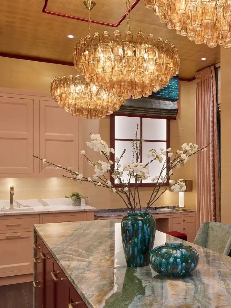 A tall glossy teal ceramic vase with textured surface on marble countertop beside amber glass ceiling chandelier