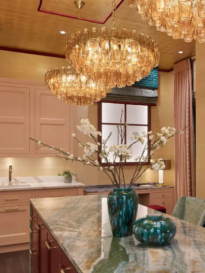 A tall glossy teal ceramic vase with textured surface on marble countertop beside amber glass ceiling chandelier