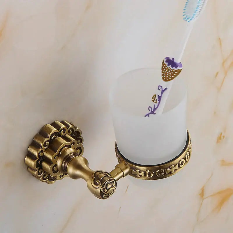 DecorBites antique brass wall mounted toothbrush holder with ornate scrollwork holding a frosted white glass cup