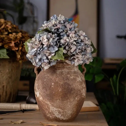 A rustic ceramic vase with decorbites artificial hydrangea silk flower arrangement in soft blue and white hues