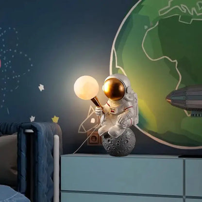 DecorBites™ astronaut led table lamp in white resin with gold helmet seated on textured gray moon surface holding glowing lamp