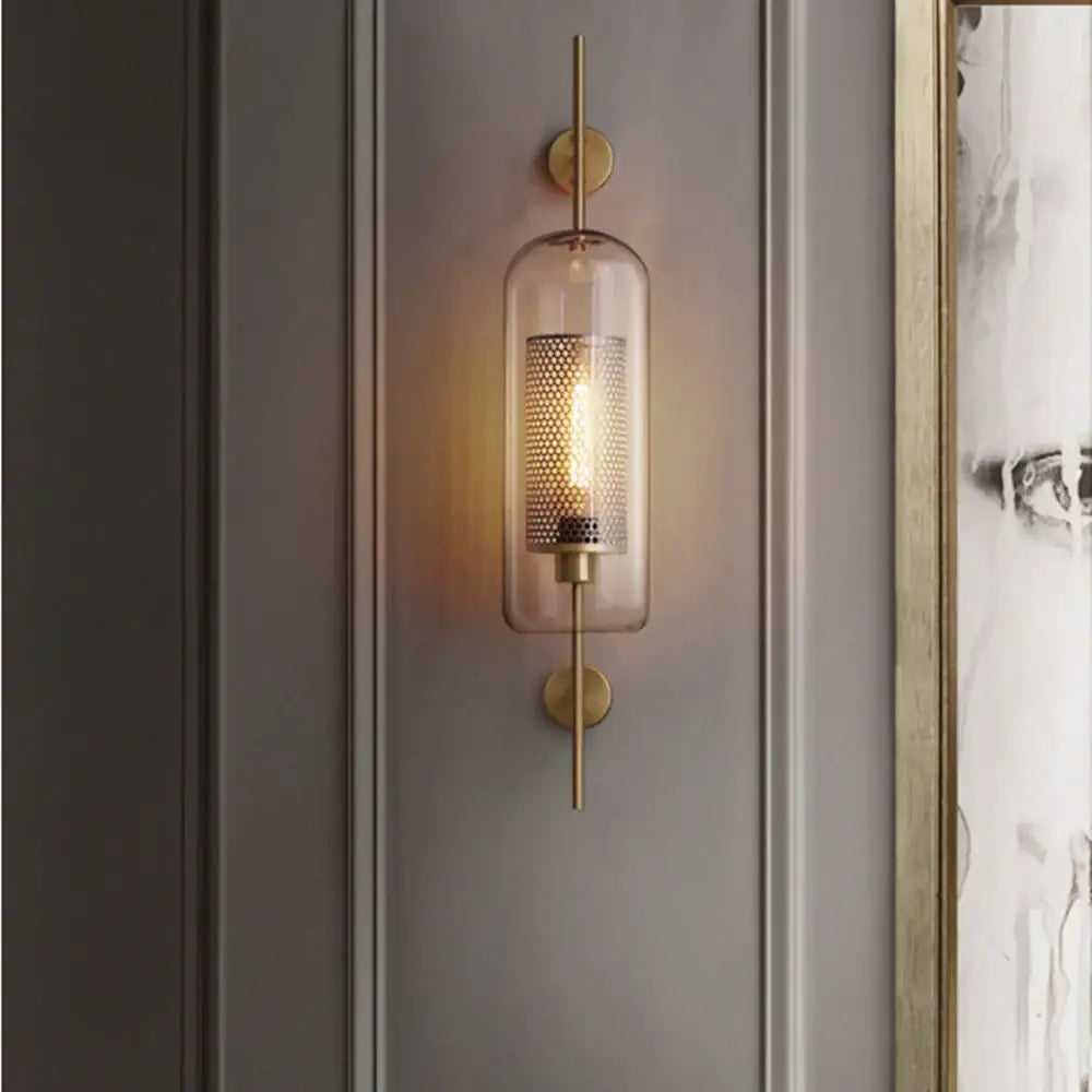 A modern brass-finished chiswick glass wall light with clear glass cylinder and perforated metal mesh for home décor