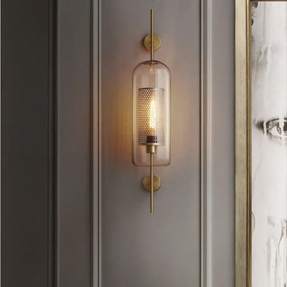 A modern brass-finished chiswick glass wall light with clear glass cylinder and perforated metal mesh for home décor