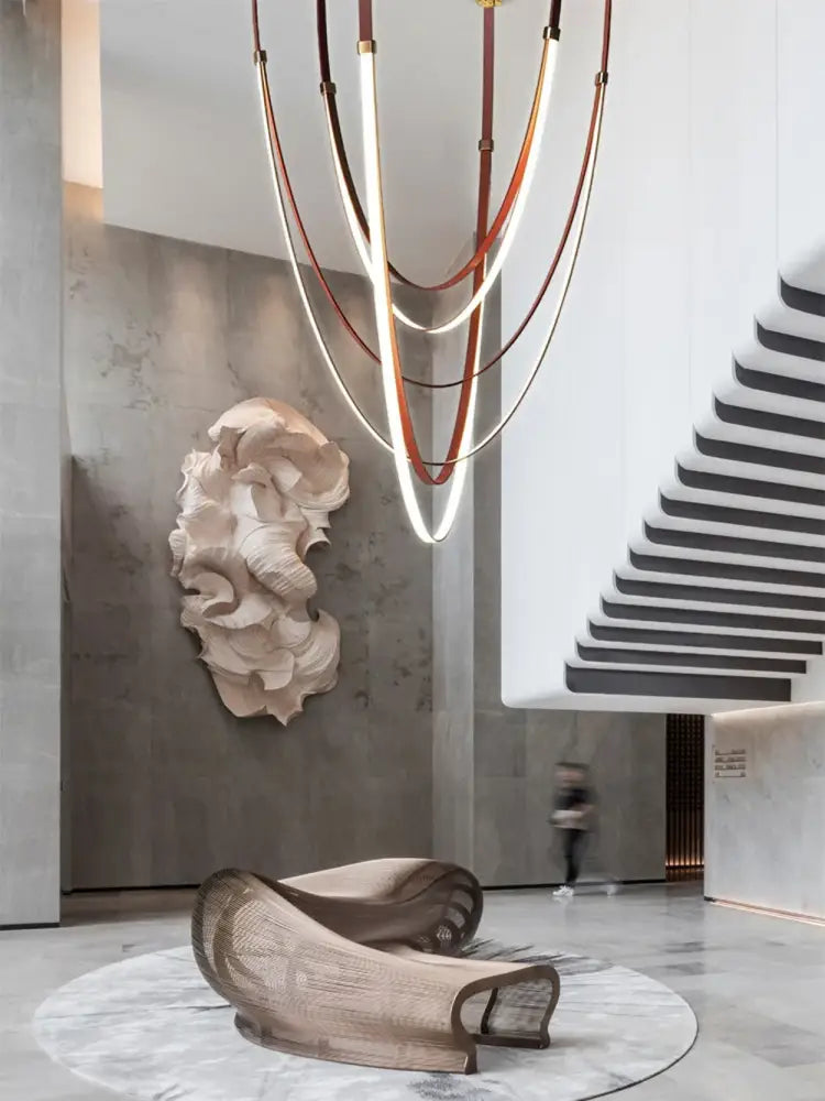 A sculptural beige lounge chair with woven mesh and curved metal frame displayed with a flexible leather belt chandelier