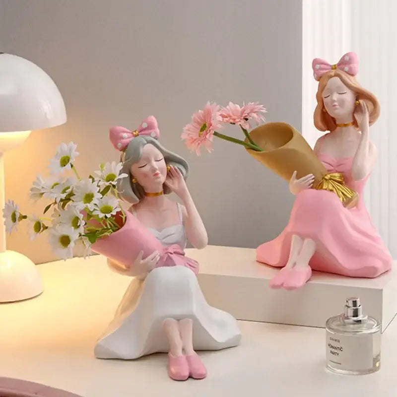 Two pink and white resin decorbites girl statues holding paper-wrapped flower bouquets as vase ornaments