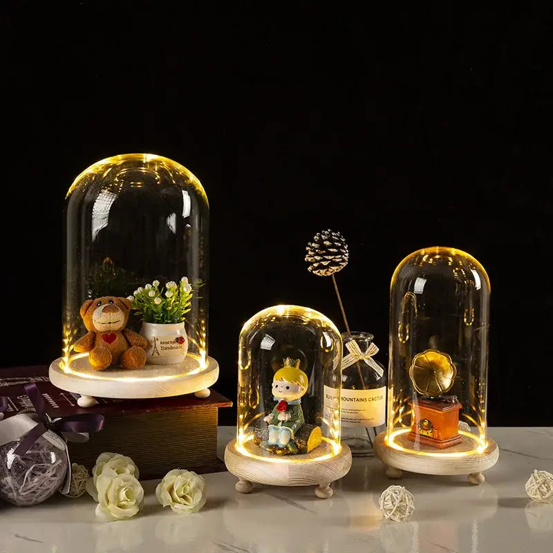 Three glass bell jar terrariums with warm LED lighting showcasing miniature scenes on wooden bases by DecorBites