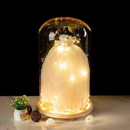 A glowing white wedding dress displayed under a clear glass bell jar terrarium with warm led fairy lights wrapped around it