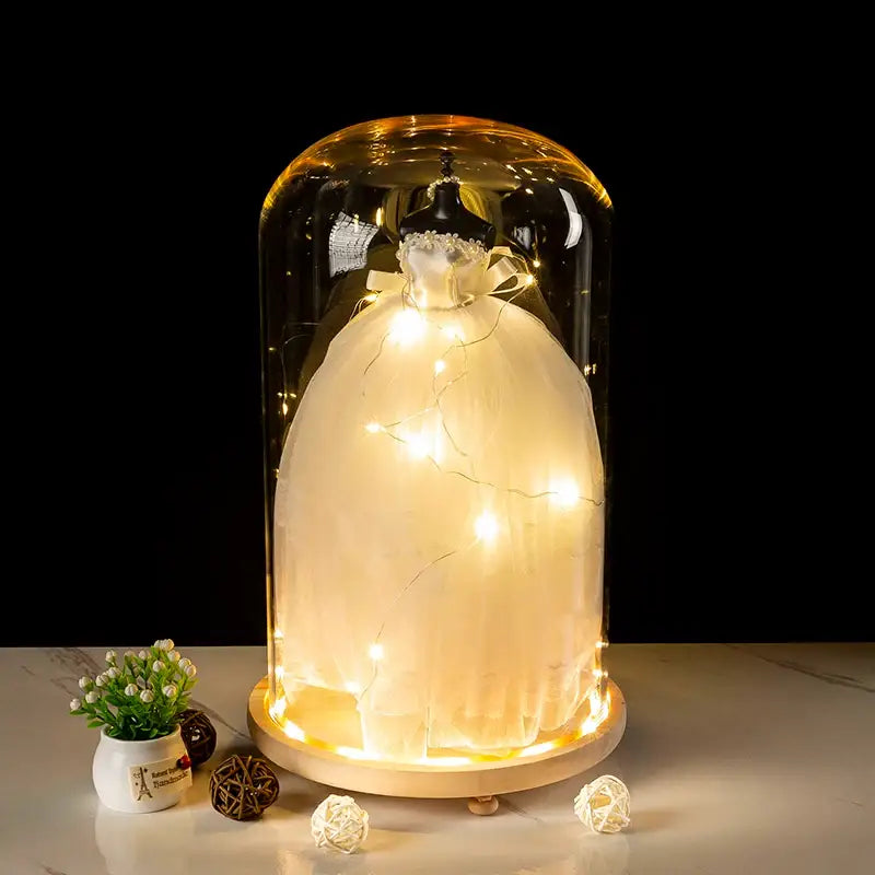 A glowing white wedding dress displayed under a clear glass bell jar terrarium with warm led fairy lights wrapped around it