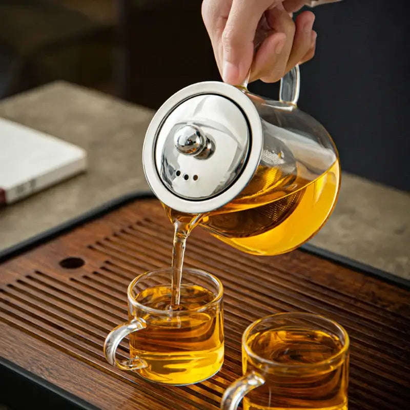 Decorbites glass tea jug set with clear glass teapot, stainless steel lid, and pouring tea into matching transparent cups