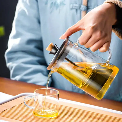 DecorBites glass tea pot with stainless steel lid and wooden handle pouring golden tea into clear glass mug