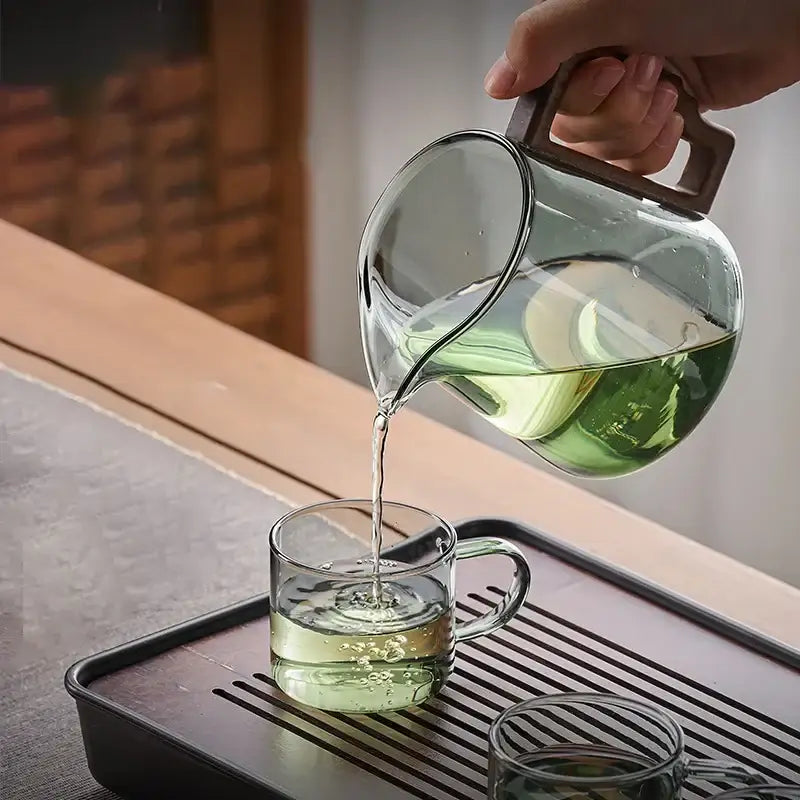 Clear glass decorbites glass teapot with dark brown handle pours green tea into matching glass cup with filter