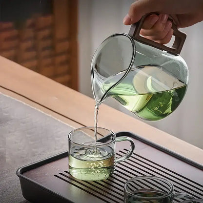 Clear glass decorbites glass teapot with dark brown handle pours green tea into matching glass cup with filter