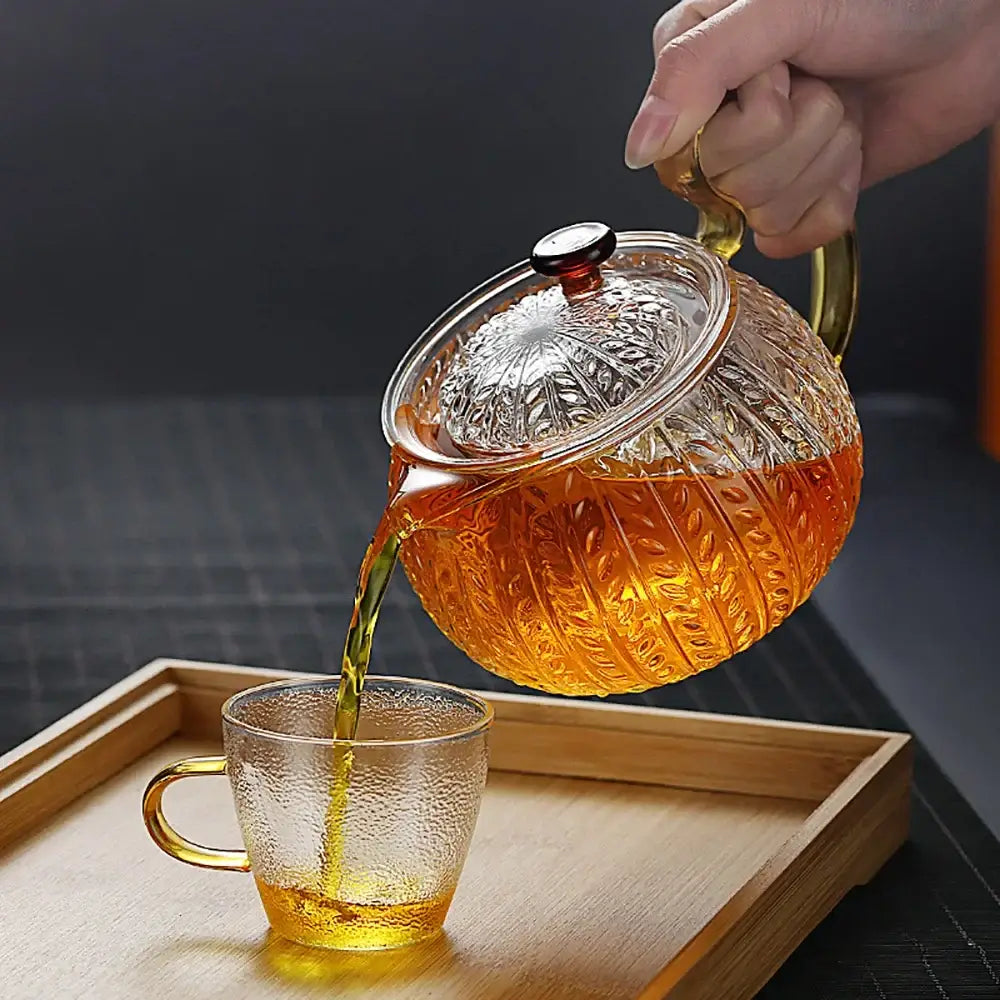 A decorbites glass teapot with leaf etching and gold handle pouring amber tea into a matching glass cup