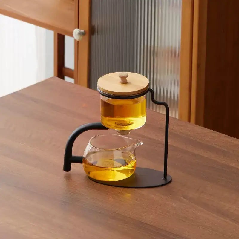 DecorBites glass teapot set with bamboo lid and black metal stand featuring six clear cups filled with golden tea