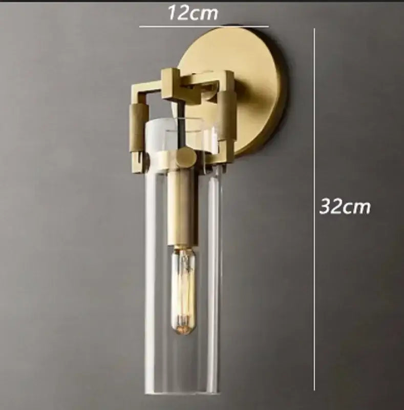 A modern brass-finished decorbites glass wall lamp with clear glass cylinder and exposed filament bulb for bedroom or dining room