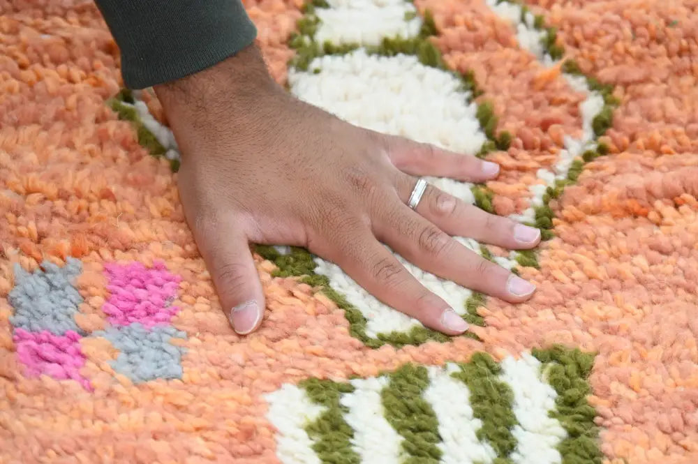 A hand rests on a vibrant handmade traditional azilal carpet with bold orange white green and pink geometric patterns