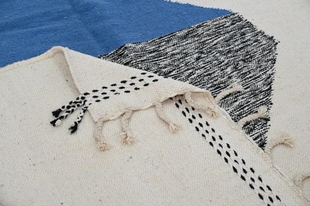 Cream-colored azilal rug with black dotted fringe and bold blue and black geometric pattern showcasing moroccan craftsmanship