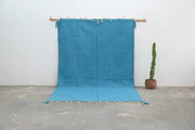 A vibrant turquoise handmade azilal rug 306x200cm with fringed edges and tassels displayed on a wooden rod