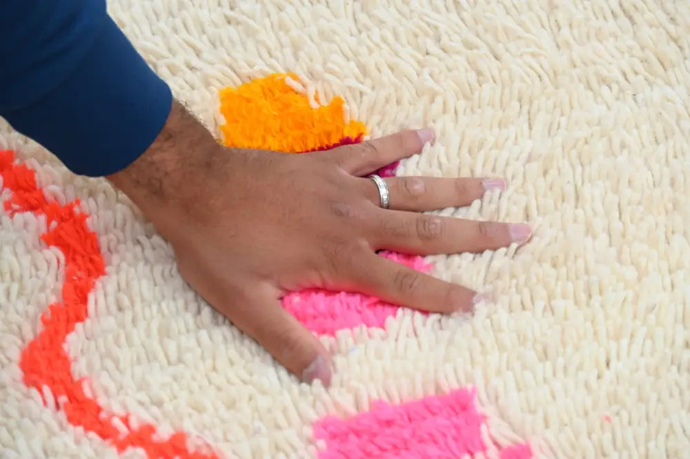 A hand with a silver ring rests on a cream handmade moroccan azilal rug with vibrant orange and pink geometric patterns