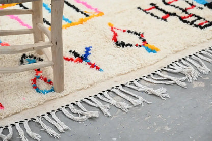 Cream-colored handwoven moroccan azilal rug with bold red blue black yellow geometric patterns and long white fringe edge