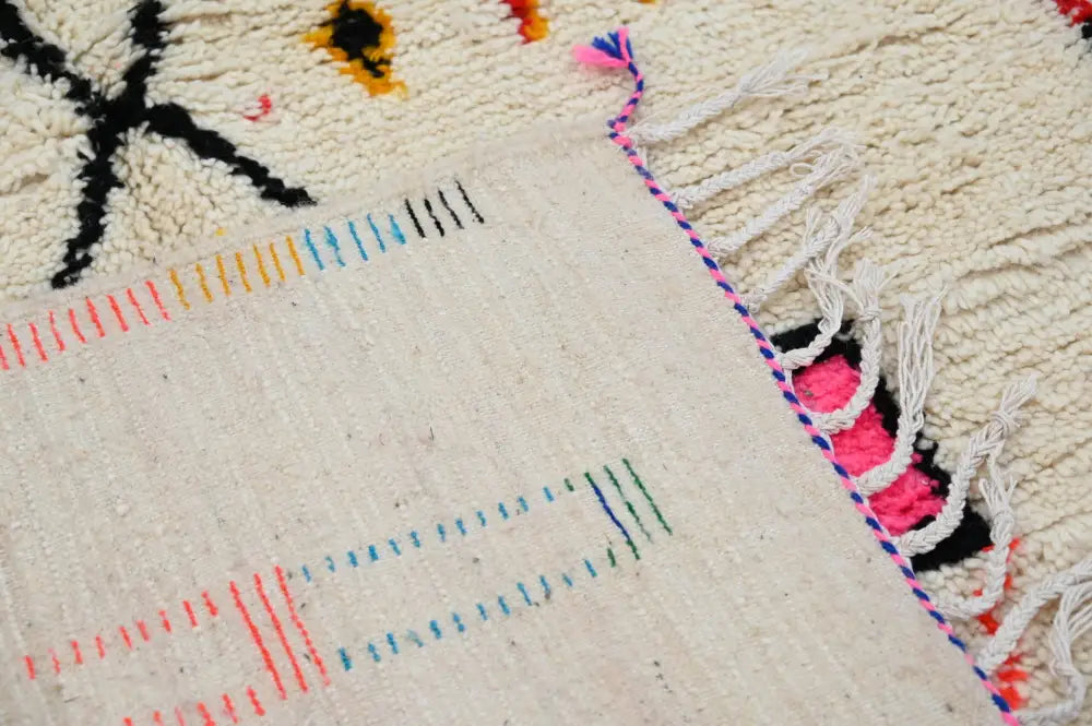 Cream-colored decorbites handwoven moroccan azilal rug with intricate black orange blue pink geometric patterns and fringed edge