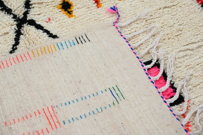 Cream-colored decorbites handwoven moroccan azilal rug with intricate black orange blue pink geometric patterns and fringed edge