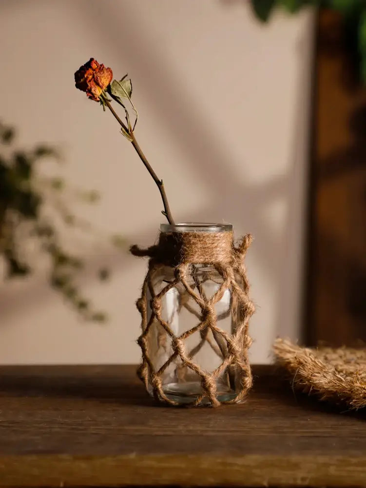 A clear hanging glass vase with hemp rope and woven jute wrap holding a single dried orange rose for rustic decor