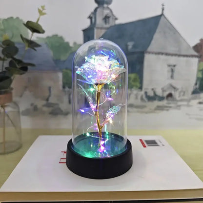 A glowing iridescent eternal rose light with a golden stem inside a clear glass dome on a black base by decorbites