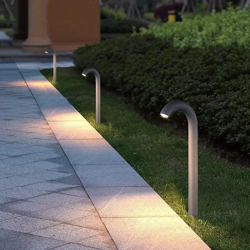 Modern curved decorbites led outdoor aluminum bollard light with sleek metallic finish emitting warm light along stone path