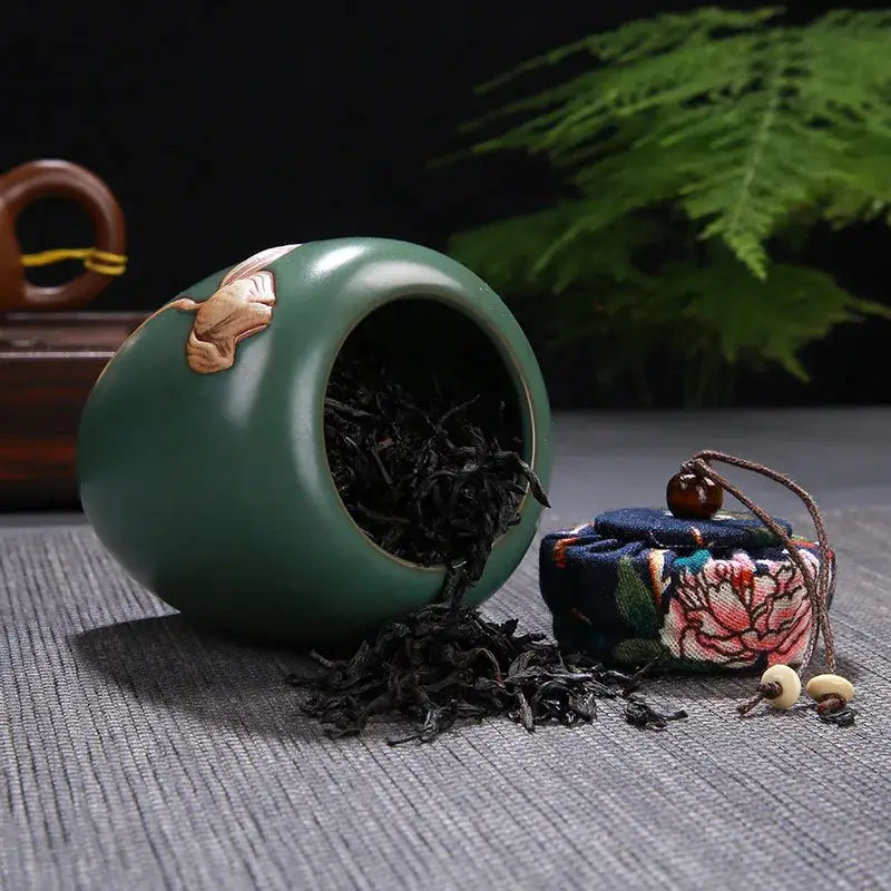 A glossy teal ceramic tea pot storage jar with gold lotus pattern spilling dark loose-leaf tea on surface