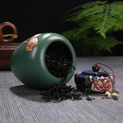 A glossy teal ceramic tea pot storage jar with gold lotus pattern spilling dark loose-leaf tea on surface