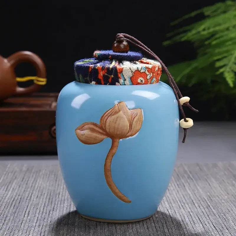 A light blue ceramic pot storage jar with hand-painted lotus pattern and fabric lid secured by brown cord for tea pot storage