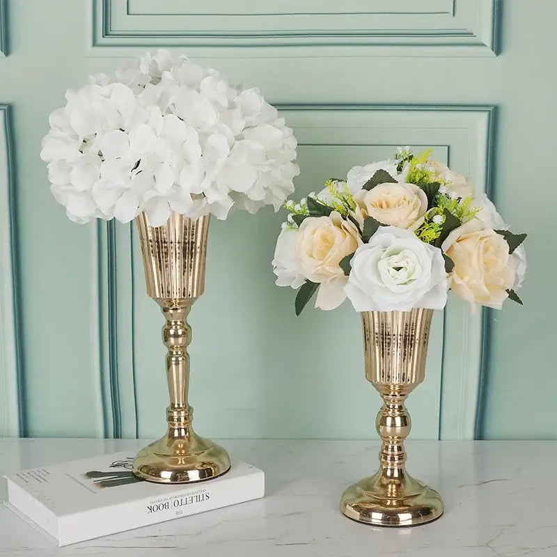 Elegant gold-toned metal trumpet vase with fluted pedestal design holding artificial white hydrangeas and cream roses