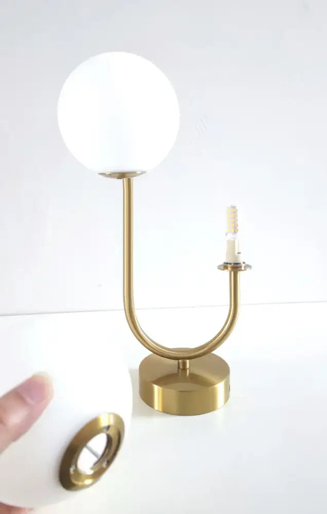 A sleek gold-toned table lamp with a frosted white spherical glass shade and curved stem for modern decor