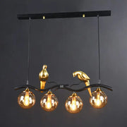 Gold-colored bird figurines perch on a black wrought iron branch holding amber glass globes in a decorbites island pendant light