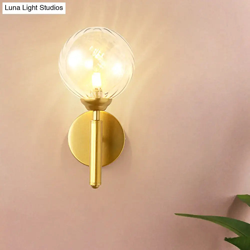 Modern brass wall lamp fixture with gold-toned cylindrical stem and clear textured glass globe for bedroom use