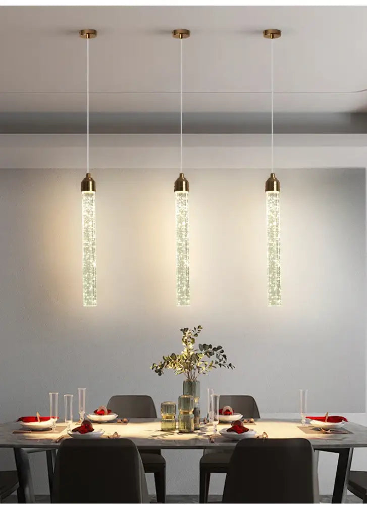 Three elegant gold-toned crystal pendant lights with sparkling glass tubes hanging above a dining table