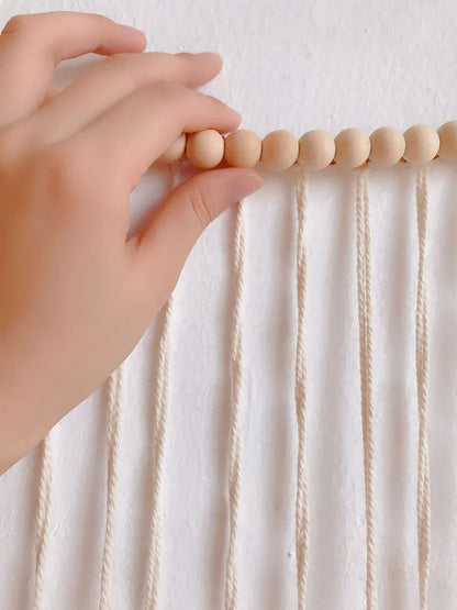 A hand gently holds light beige wooden beads on cream macrame cords in moroccan bohemian wall decor tapestry