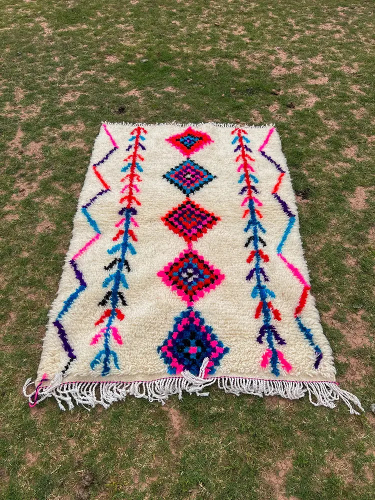 A vibrant cream-colored moroccan handmade azilal rug with bold pink blue red and purple geometric patterns and fringed edges