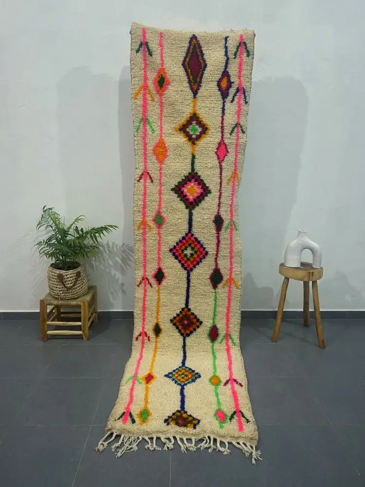 A vibrant cream-colored moroccan handmade azilal rug with bold pink orange green and purple geometric patterns and fringed edge