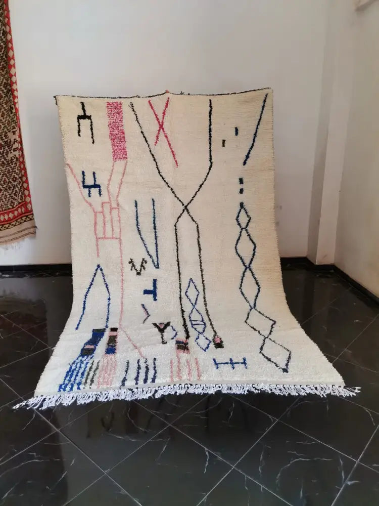 Cream-colored moroccan handmade azilal berber carpet with black pink and blue geometric patterns and fringed edges supporting sustainable practices